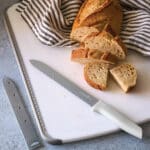 Pampered Chef beschichtetes Brotmesser mit hellgrauem Griff und grauer Antihaftbeschichtung, auf weißem Schneidebrett mit geschnittenem Brot und gestreiftem Geschirrtuch
